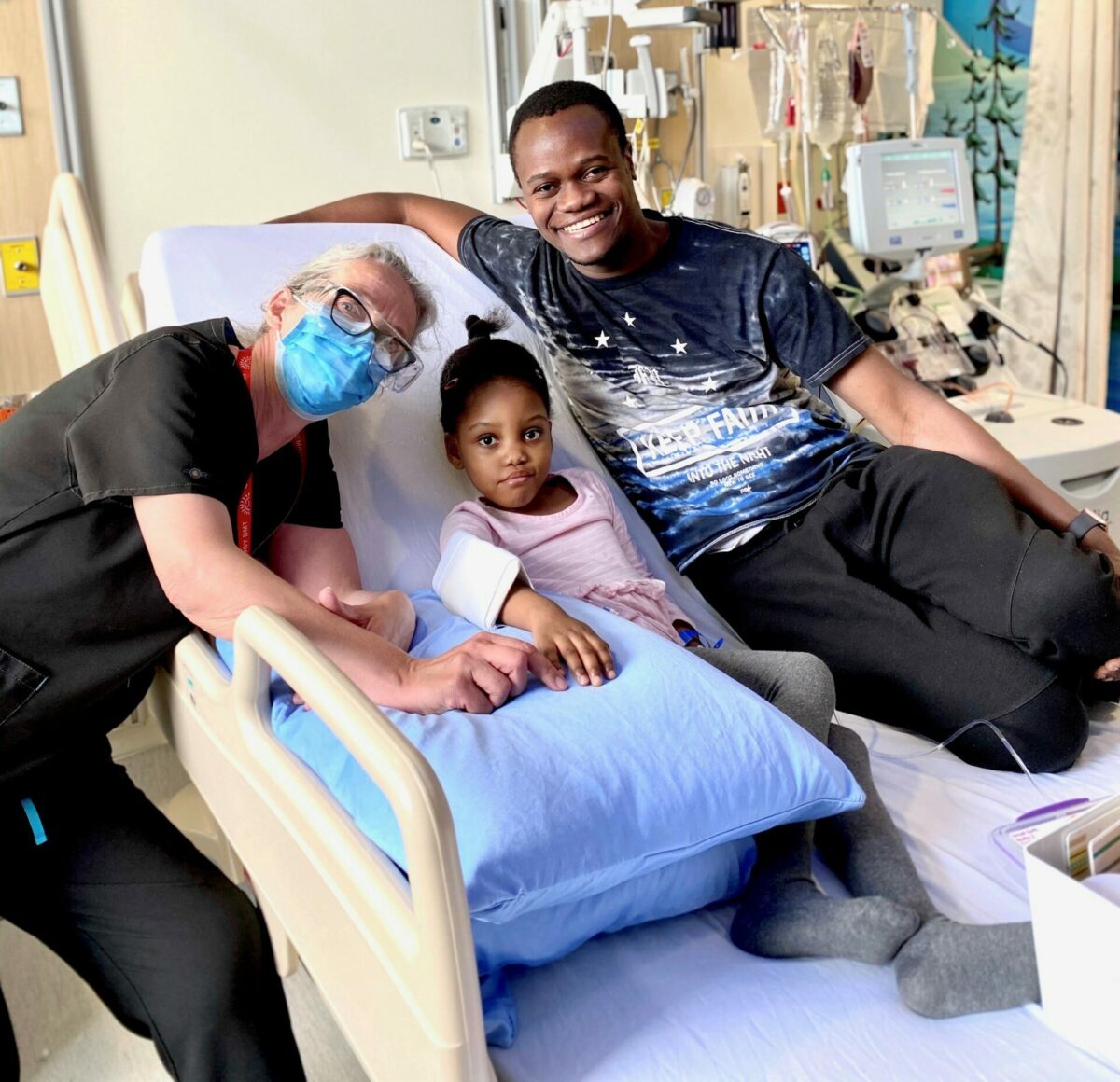 Young patient sits on hospital bed with father and attending nurse smiling on either side