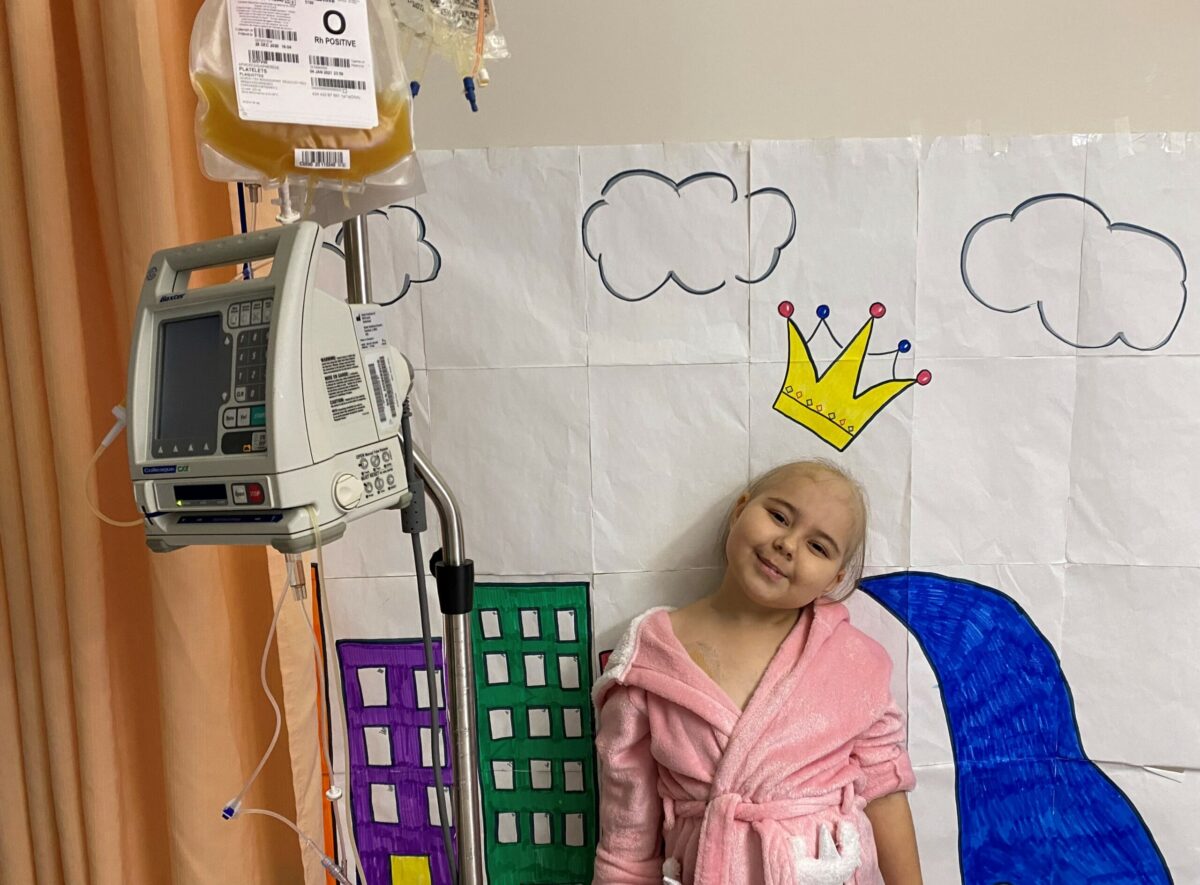 Young patient with cancer smiles while standing next to a bag of donated blood platelets in a hospital