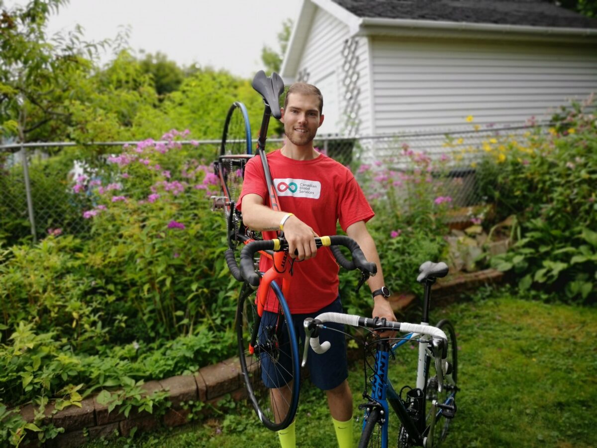 Young man stands smiling in a garden with a bike on his shoulder