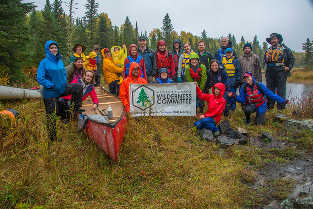 A group of people gather on a grassy slope in their raingear, canoe and Wilderness Committee sign in the fore.