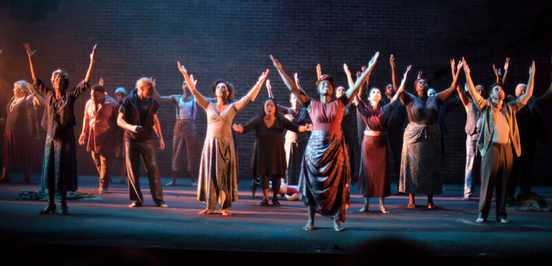 Two dozen actors on an empty stage with their hands raised towards the sky Production: The Adventures of a Black Girl in Search of God by Playwright Djanet Sears