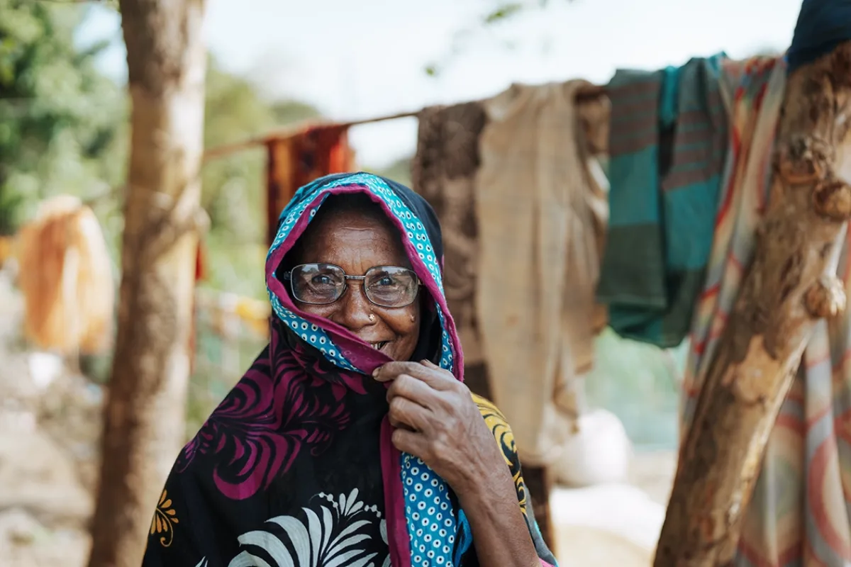 A woman wearing glasses peers out, smiling, from behind a sari that is pulled over her head.