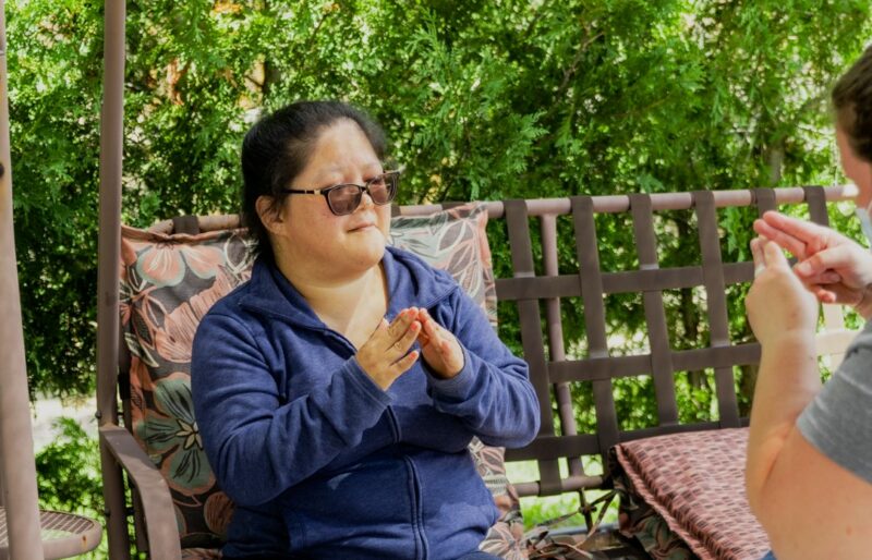 Woman with dark hair and sunglasses is using sign language to communicate with her intervenor.