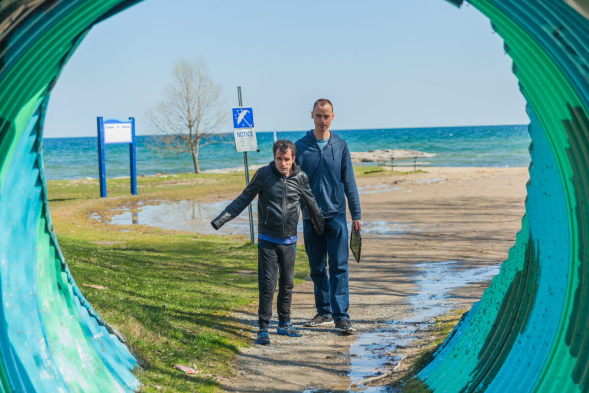 Man with dark hair and black jacket walking through a colourful tunnel with the beach and lake behind him and his intervenor
