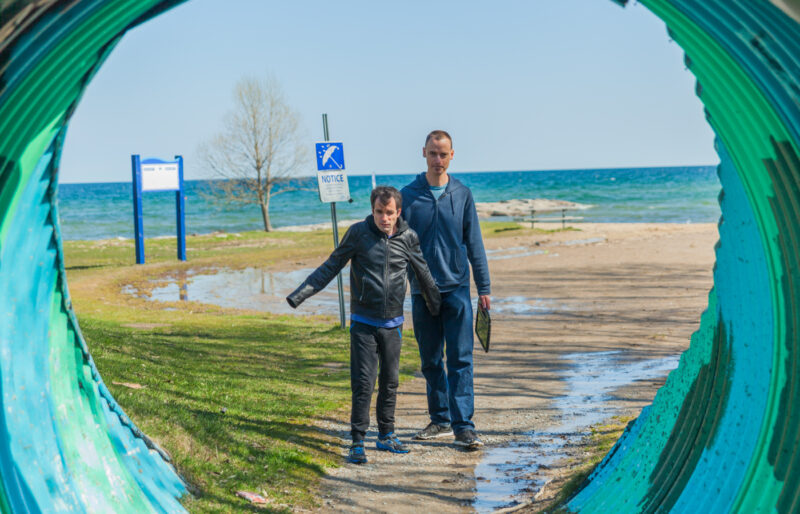 Man with dark hair and black jacket walking through a colourful tunnel with the beach and lake behind him and his intervenor