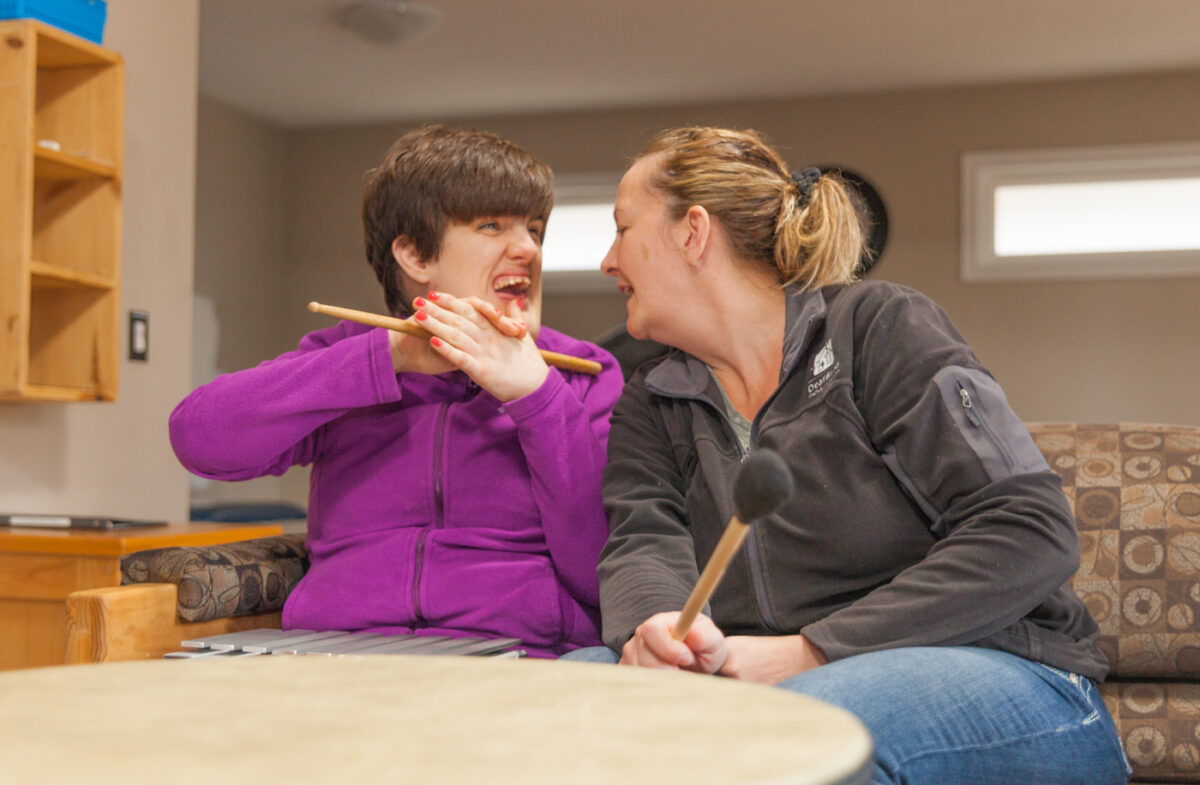 Woman with short dark hair and purple jacket holding a drum stick in front of a drum laughing with her intervenor