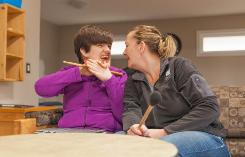 Woman with short dark hair and purple jacket holding a drum stick in front of a drum laughing with her intervenor