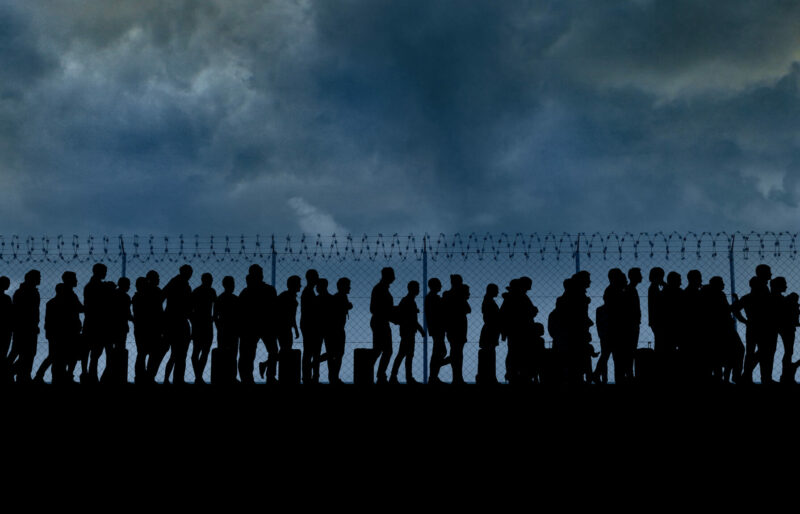 Line-up of migrants near the USA border against a fence with barbed wire.