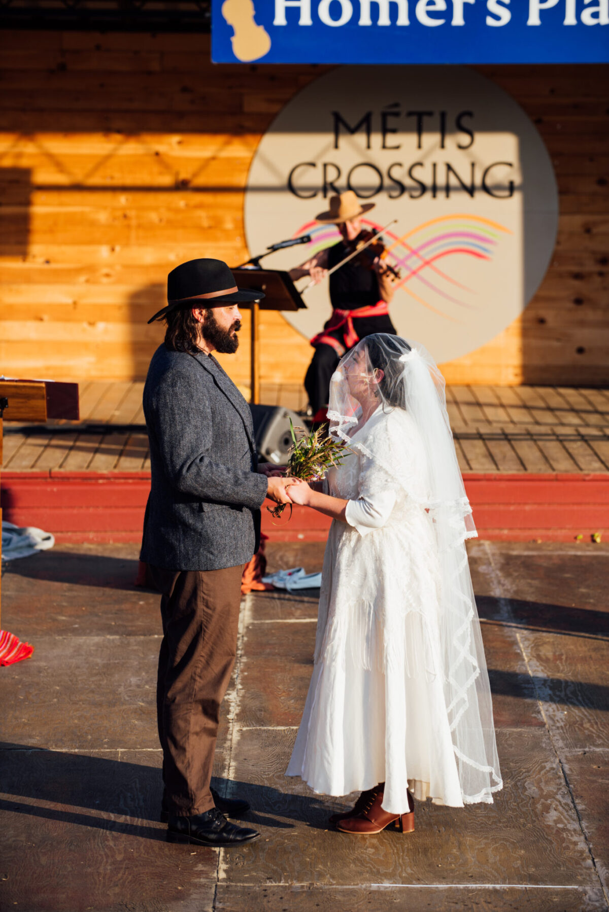 Two actors and a musician are standing on an outdoor stage on a sunny day. The actors are holding hands and facing each other. One actor is wearing a white wedding dress and the other actor has on a black cowboy hat, grey suit jacket and brown pants. A musician plays the fiddle in the background in front of a sign that reads Metis Crossing.