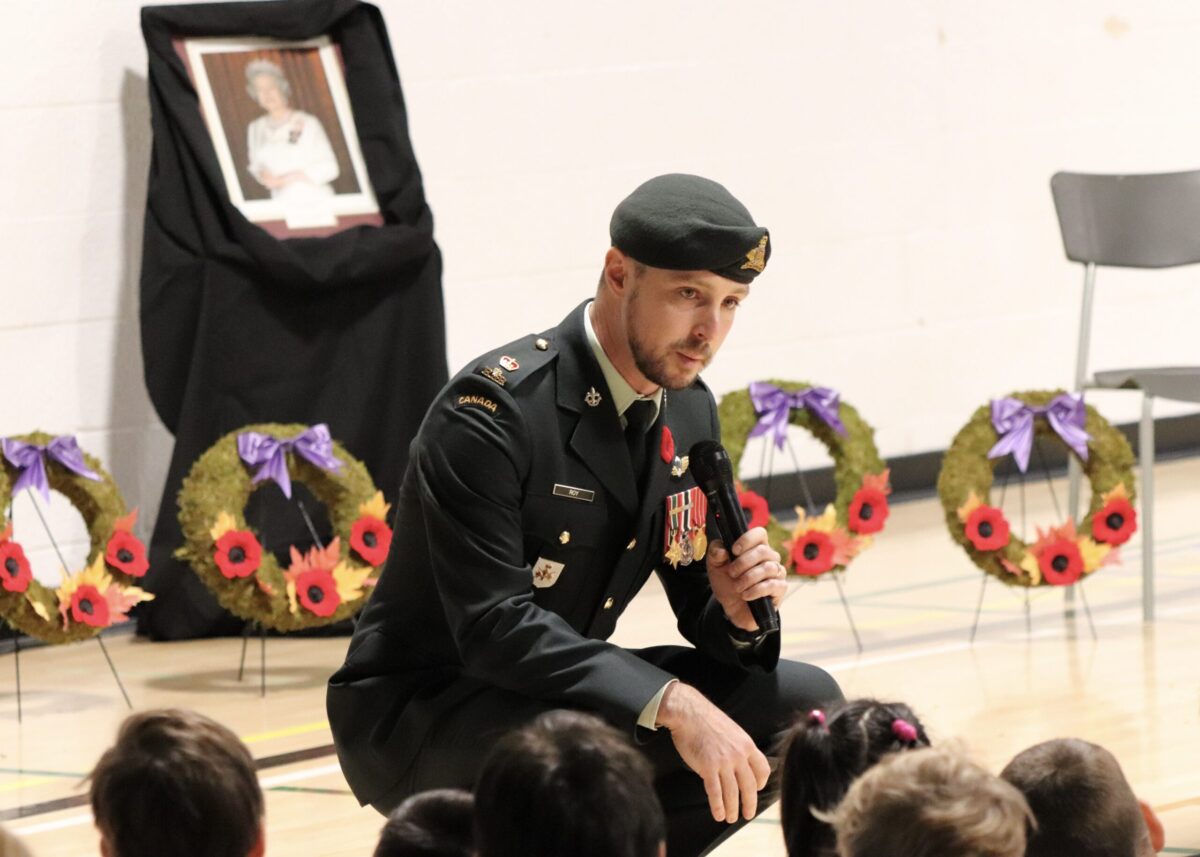 A uniformed officer holding a microphone speaks to a gymnasium of students.