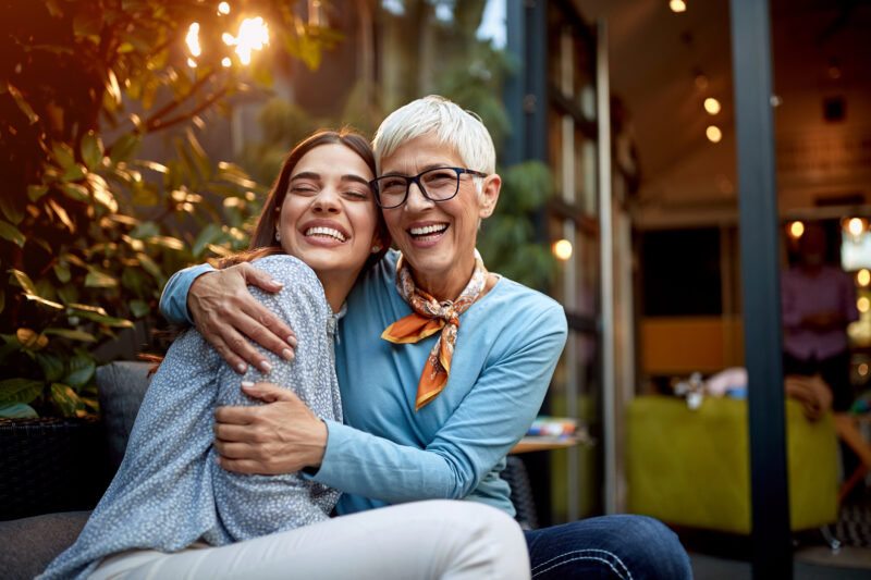 A photo of a senior mother and adult daughter sitting down hugging each other and smiling.