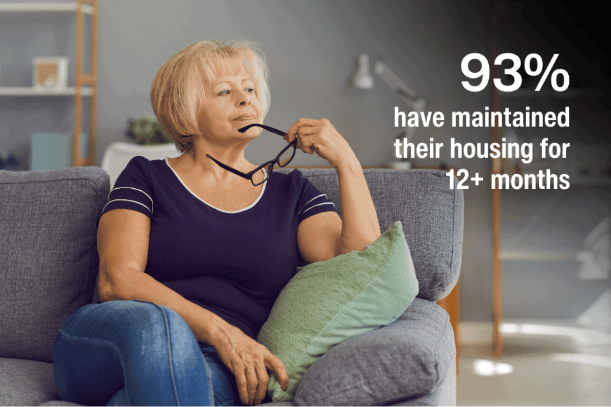 middle-aged woman wearing navy shirt and jeans sitting on sofa holding eyeglasses. TEXT SAYS: 93% have maintained their housing for 12+ months