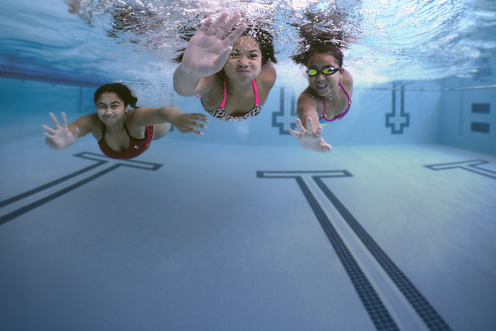 Three kids aged between 5 and 10 are swimming in a pool. The photo is shot from underwater looking up at them.