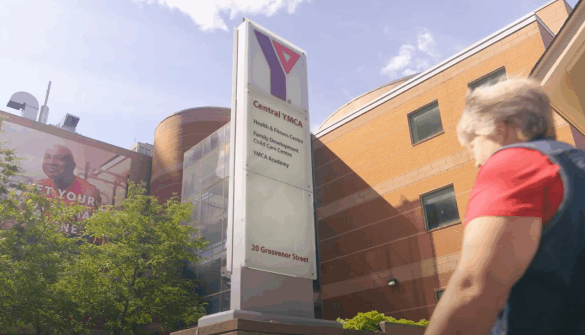 A senior woman approaches a red brick 2 story building. In front there is a tall sign saying it is the Central Y