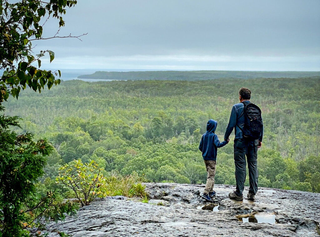 Father and son standing on a rock outcrop overlooking a forest and lake along the Bruce Trail, symbolizing protecting natural spaces for future generations.