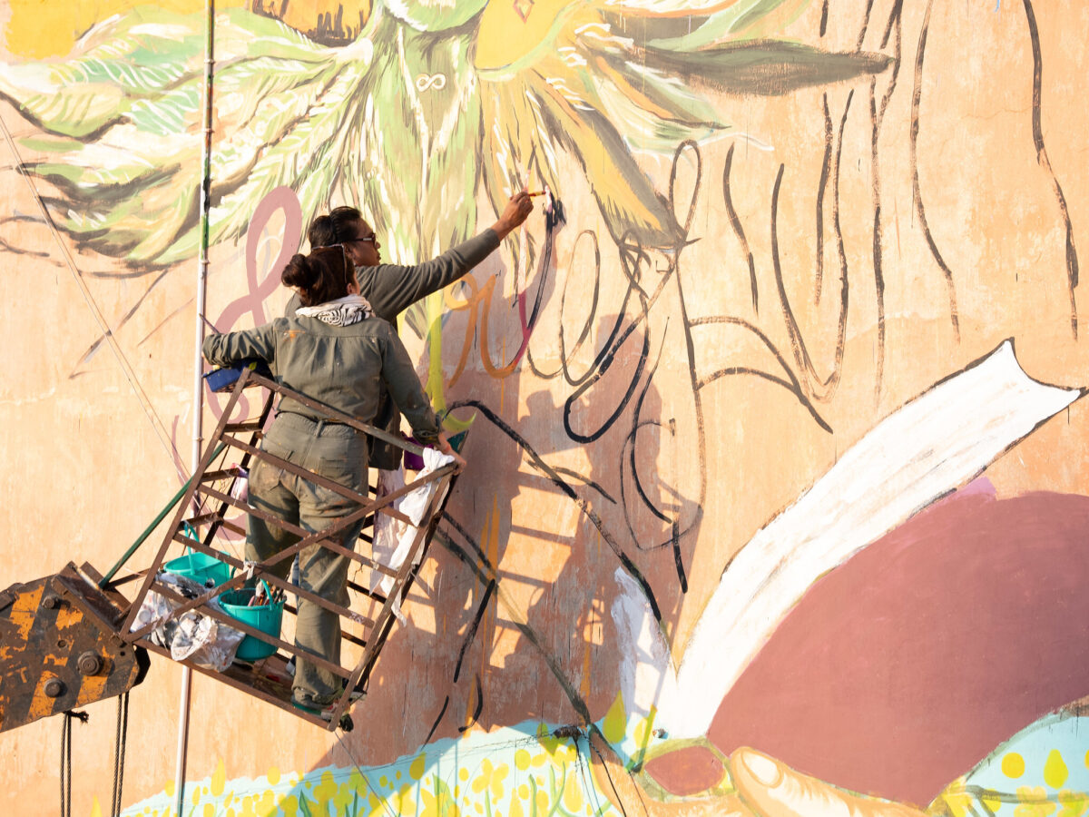 Two women stand on scaffolding to paint a mural on the side of a building. The part they are paying says 