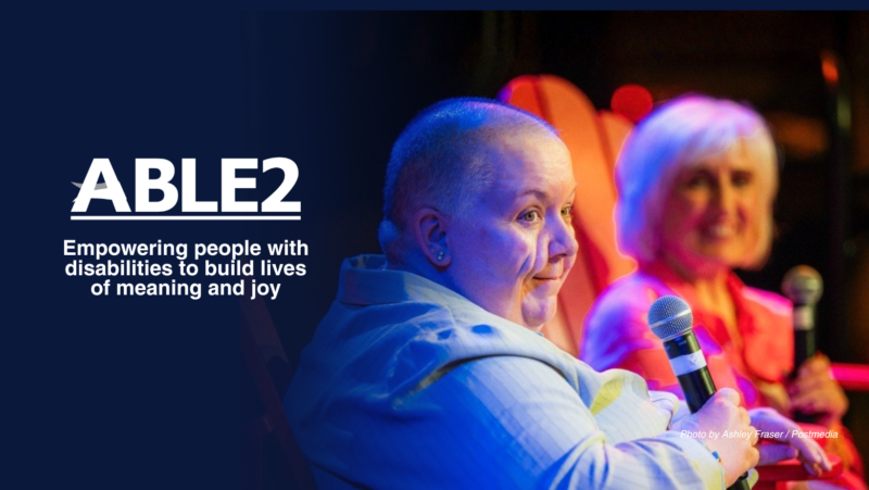 ABLE2 white logo. Image of a person with disability and her volunteer Ally speaking to an audience. Text displayed : ABLE2 empowering people with disabilities to build lives of meaning and joy